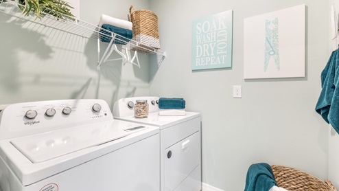 Inside of the Cali model laundry room. Room contains a white washer and dryer side by side. There is also a shelf above the washer and dryer with hangers on it.