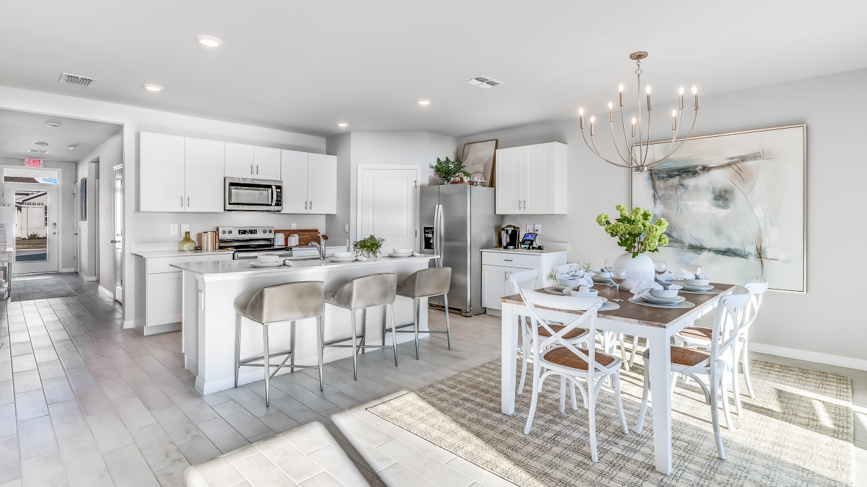 Inside the Cali Model kitchen and Dining room. You can see the entrance hallway from the door. This home includes tiles flooring, stainless steel appliances and white cabinetry with silver fixtures. The Dining space is well lit from the patio doors.