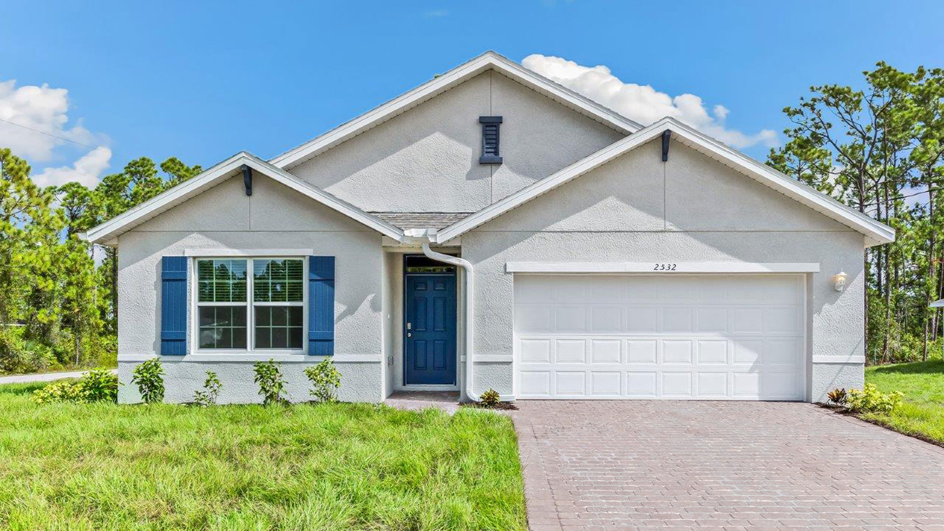 Outside of the Cali D model. Grey home with a two car garage, one window with shutters, and front door. Mixed pavers driveway.