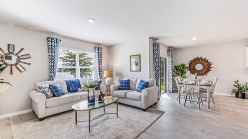 Inside of the Cali model living room. Grey wood tile floors and off-white walls. Room contains two small couches and a coffee table. The dining room can be seen in the background as well as the door to the outdoor patio. Room also contains a large window.