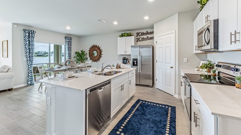 Inside of the Cali model kitchen. This angle shows the dishwasher on the inside of the kitchen island as well as the other stainless-steel appliances such as a refrigerator, stove, and microwave. In the background the dining room and back patio can be seen.