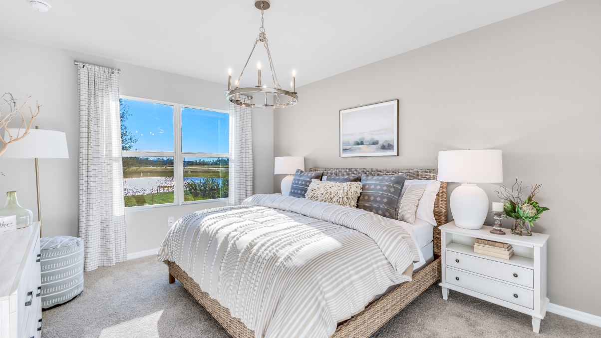 The Master Bedroom is carpeted with grey painted walls. There is a window with a view of the back of the home. There are side tables on either side of the freshly made bed. There is a chandelier above the bed as well.