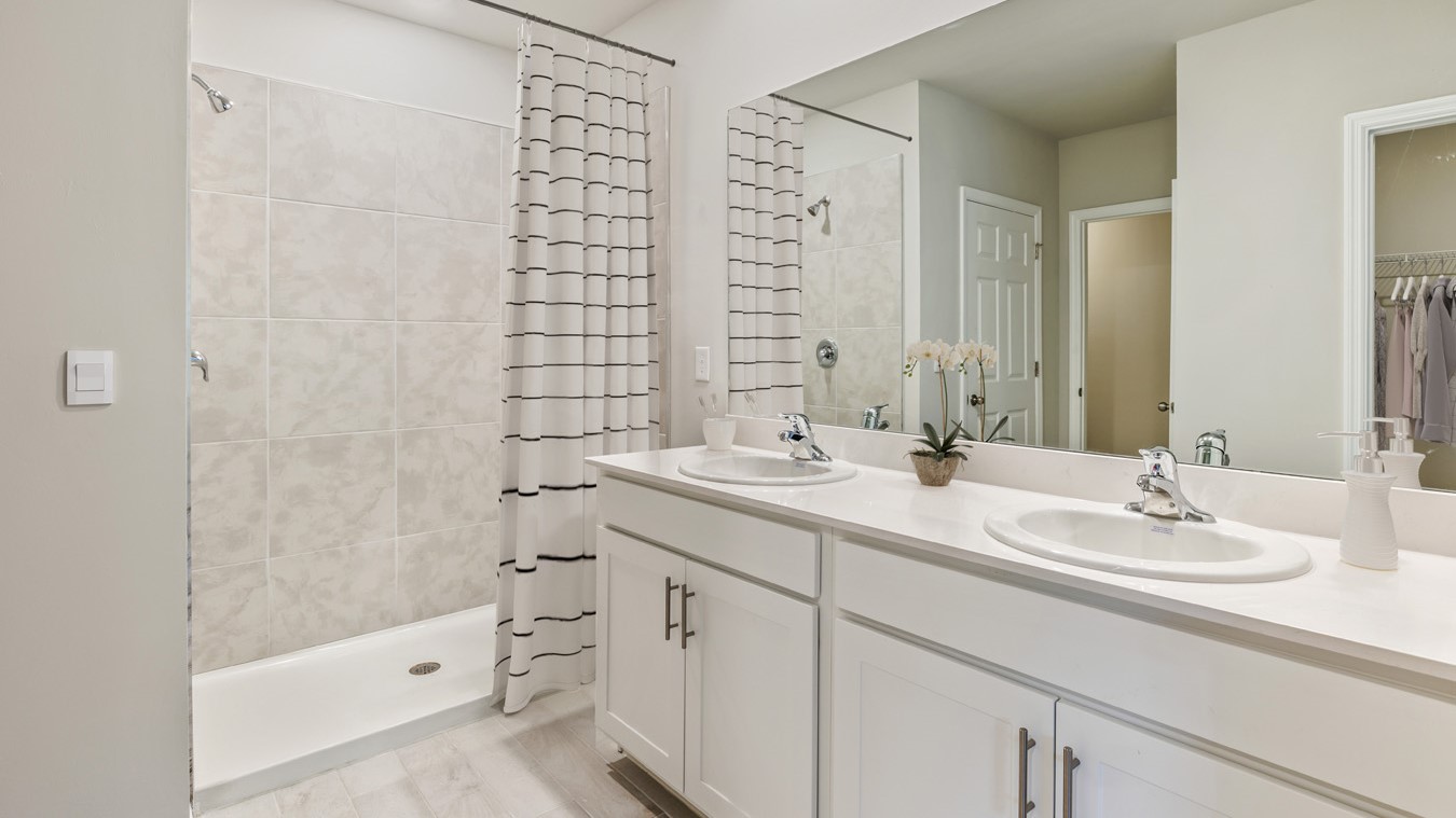 Inside of the Madison bathroom one. Double vanity with white countertops, white cabinets, silver hardware. White wood tile flooring off-white walls. Shower with grey tile and white flooring. Closet connected to bathroom.