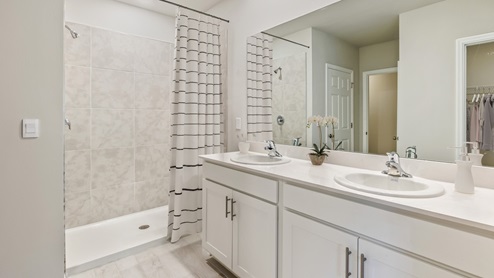 Inside of the Madison bathroom one. Double vanity with white countertops, white cabinets, silver hardware. White wood tile flooring off-white walls. Shower with grey tile and white flooring. Closet connected to bathroom.