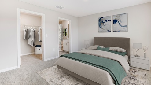 Inside of the Boardwalk bedroom one. Off-white walls with beige carpet. Room contains a walk-in closet, connected bathroom, bed with grey sheets, and small nightstands on either side of bed.
