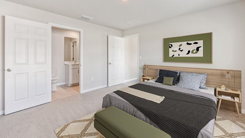 Inside of the Freeport bedroom. White carpet with creme walls. Bedroom has a connected bathroom. Room contains a large bed with grey sheets as well as two small nightstands on either side of bed.
