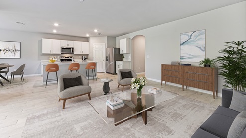 Inside of the Madison living room. Wood tile flooring with off-white walls. Room contains two armchairs with small table between them, wooden storage shelf, coffee table, and small couch. Photo demonstrates that the living room opens up to the kitchen, dining room, and hallway that connects to the front door.