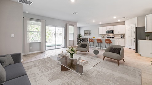 Inside of the Madison living room. This photo demonstrates that the living room also connects to the outdoor patio and has two windows on either side of the sliding glass door that leads out. Kitchen can be seen in the background to have bar-style seating, white cabinets, white countertops, and stainless-steel appliances.