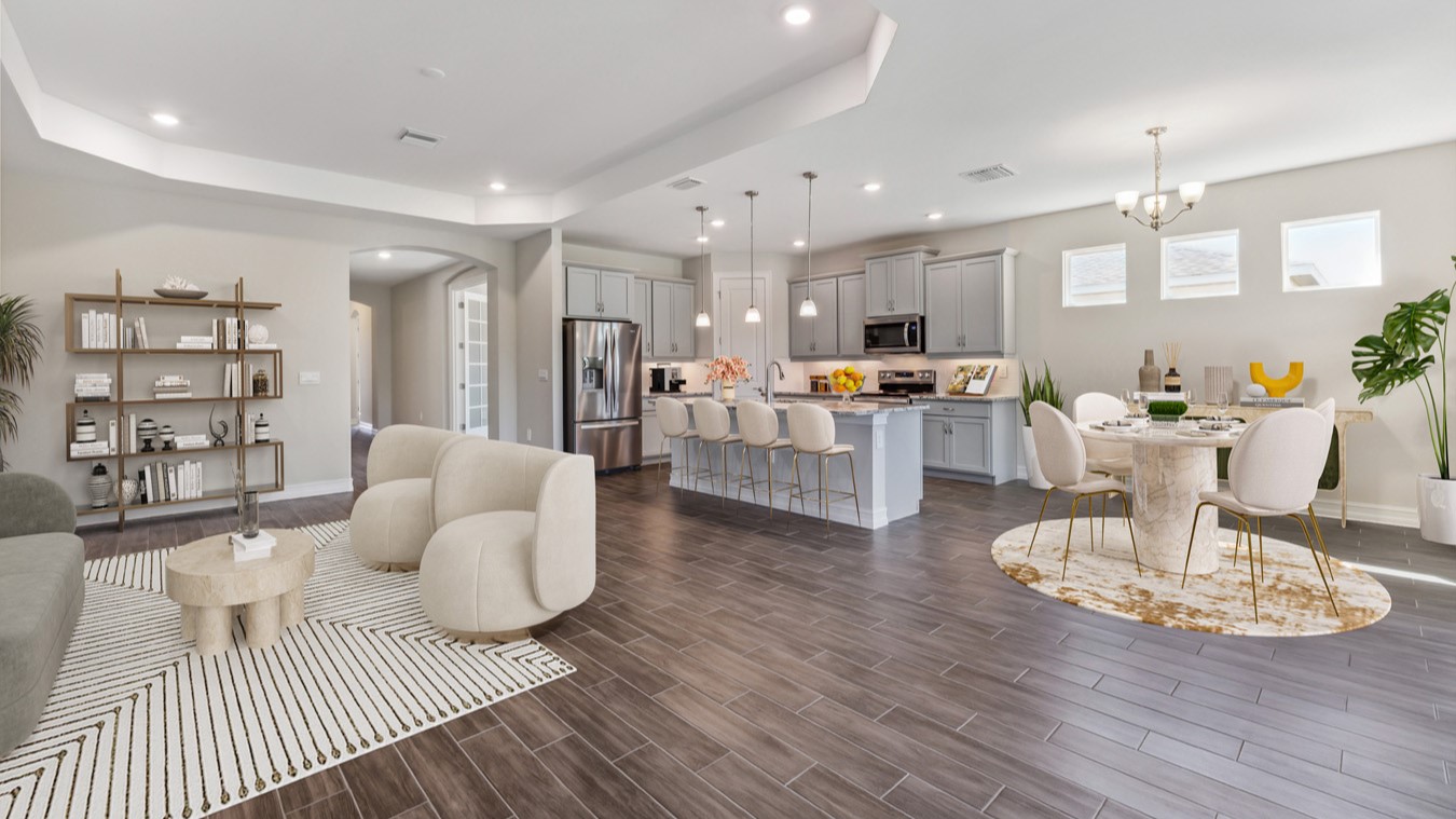 Inside of the Shelby model dining and living area. Plank tile floors with grey walls. Dining area contains a four-person dining table with hanging light fixture above it. Living area with furniture can be seen in the background as well as the hallway to the front door. Kitchen can also be seen in the background with grey cabinets and white countertops with silver hardware. Kitchen contains kitchen island with bar-style seating and stainless-steel appliances.