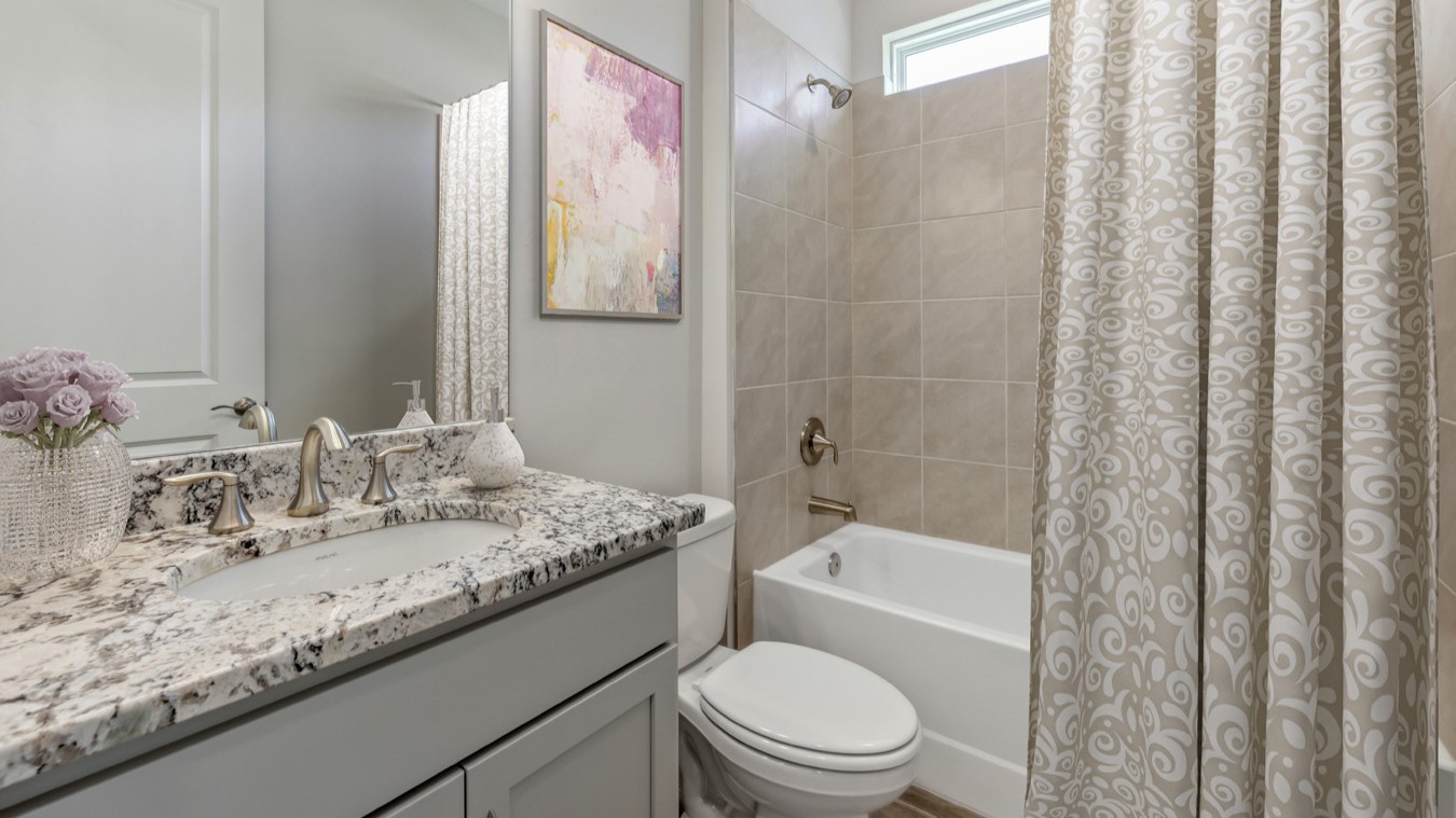 Inside of the Shelby model bathroom. Plank tile flooring and grey walls. Grey and brown countertops and grey cabinets with silver hardware. Shower-tub combo has grey tiles and white tub. White toilet between vanity and shower-tub combo.