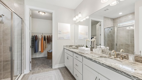 Inside the Clifton primary bathroom with dual sinks and shower without a tub. You can see into the master closet from this angle. Cabinets are white with silver handles. Marble countertop and brown tiles in shower.