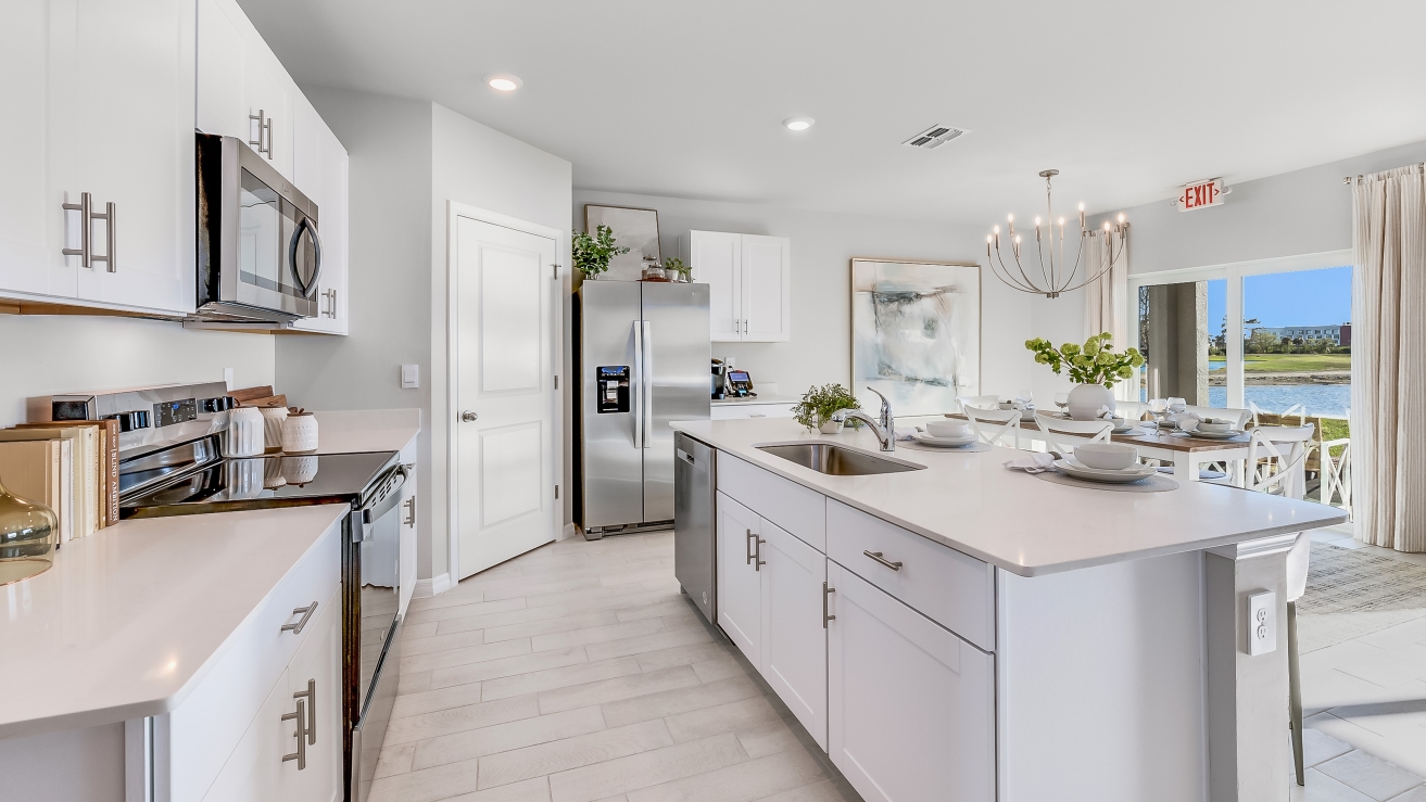 Inside the Cali Model kitchen. This home includes tiles flooring, stainless steel appliances and white cabinetry with silver fixtures. The bar is great for hosting people in the kitchen. You can see the dining room and patio from here. Theres a clear view outside.