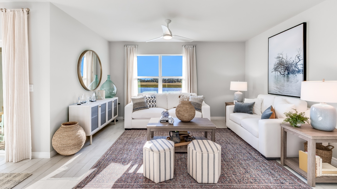 The Living room, there are two white couches, some stools, a coffee table and a chest for storage. There are wooden side tables on either side of the couch. The room is well lit with a window overlooking a view of the back.