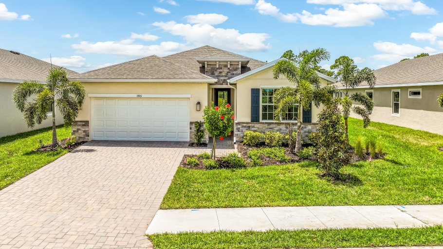 Outside of the Delray model. Yellow home with two car garage, one window with shutters, and front door. Mixed paver driveway.