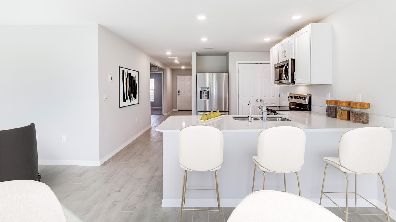 Inside of the Boardwalk kitchen. This photo shows the bar sitting available. Demonstrates how kitchen and dining area open up to the hallway leading to the front door. Kitchen contains grey wood tile floors and off-white walls. White countertops with silver hardware. White cabinets. Stainless steel refrigerator, stove, microwave, and sink.