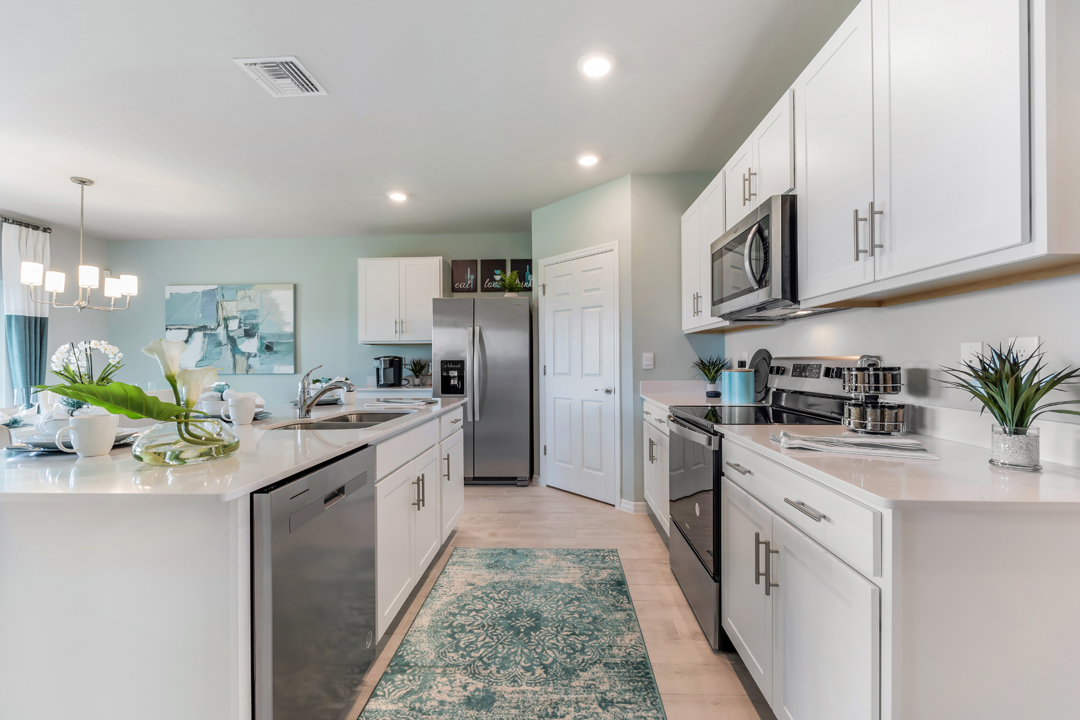 Inside of the Cali model kitchen. Wood tile floors with teal walls. Kitchen contains white countertops and cabinets with silver hardware. There is a kitchen island with bar-style seating. Kitchen appliances are stainless-steel.