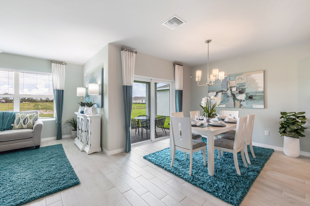 Inside of the Cali model dining area. Wood tile flooring and teal walls. Dining area contains a six-person dining table with a hanging light fixture above it. In the background a sliding glass door that leads to the back patio can be seen as well as an open area that contains the living room furniture.