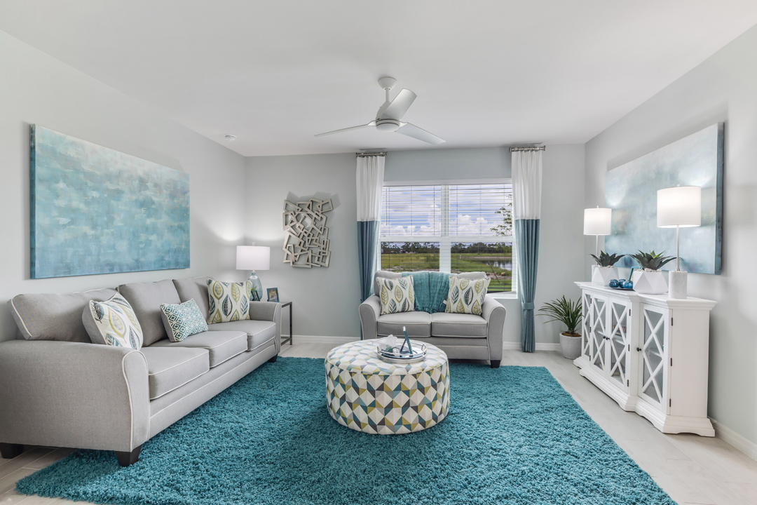 Inside of the Cali model living area. Wood tile flooring and teal walls. Living area contains two couches, an ottoman, storage shelf, and small table. Room also contains a window.