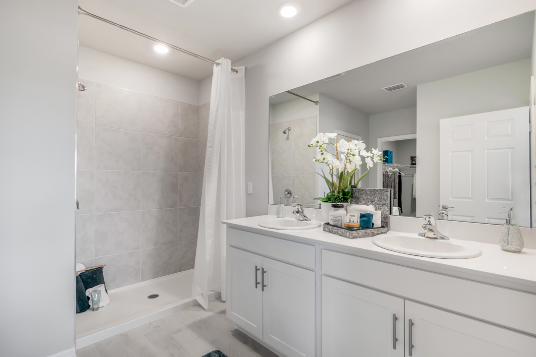 Inside of the Cali model bathroom. Shower has grey tile and silver hardware. Room contains a dual vanity with white countertops and cabinets with silver hardware. Bathroom also contains a closet.