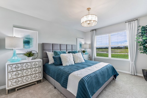 Inside of the Cali model bedroom. Crème carpet with off-white walls. Room contains a bed with nightstands on either side as well as a plant in the corner of the room.