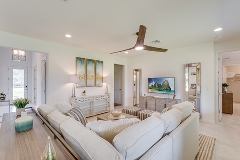 Inside of the Kellen model living room. Tile flooring and white walls. Living room has a couch, side table, two ottomen, and a coffee table. In the background you can see the front door.