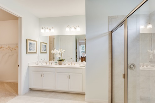 Inside of the Kellen model bathroom. Tile flooring and white walls. Double vanity and white cabinets with silver hardware. Shower has grey tile and silver hardware.