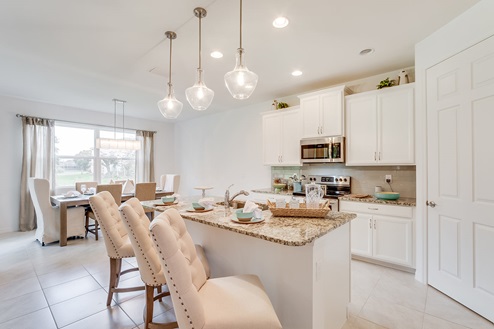 Inside of the Kellen model kitchen. Tile flooring and white walls. Kitchen has white cabinets and brown countertops with silver hardware. Kitchen island has bar-style seating. In the background the dining area can be seen.