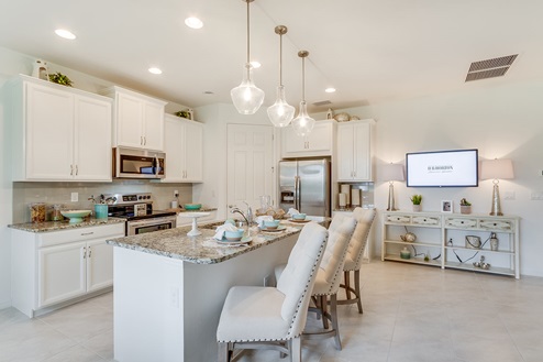 Inside of the Sage model kitchen. Tile flooring and white walls. Kitchen has white cabinets and brown countertops with silver hardware. Kitchen island has bar-style seating.