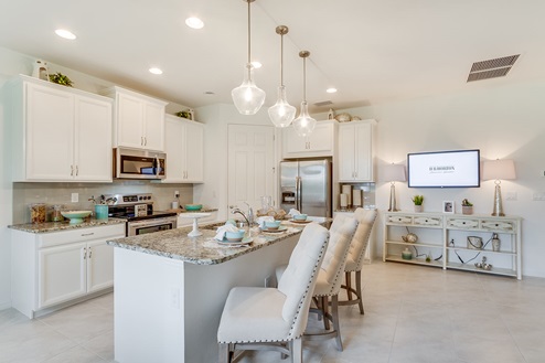 Inside of the Kellen model kitchen. Tile flooring and white walls. Kitchen has white cabinets and brown countertops with silver hardware. Kitchen island has bar-style seating.