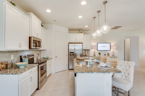 Inside of the Sage model kitchen. Tile flooring and white walls. Kitchen has white cabinets and brown countertops with silver hardware. Kitchen island has bar-style seating.