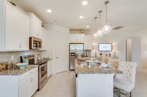Inside of the Kellen model kitchen. Tile flooring and white walls. Kitchen has white cabinets and brown countertops with silver hardware. Kitchen island has bar-style seating.