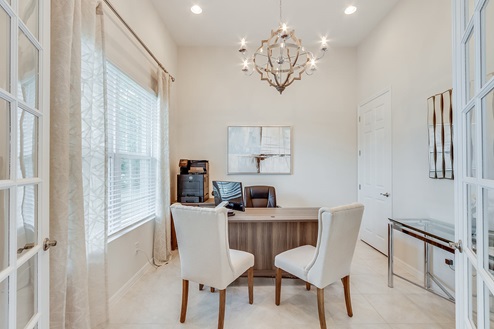 Inside of the Kellen model den. Tile flooring and white walls. Den has a desk with laptop on it, three chairs, glass desk, and small dresser with printer on it. Room also has window.