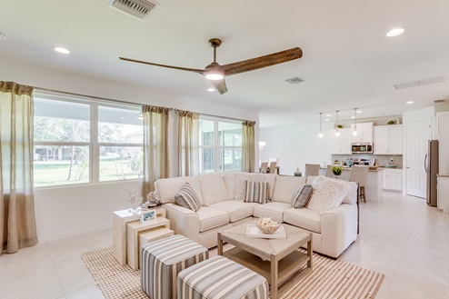 Inside of the Kellen model living room. Tile flooring and white walls. Living room has a couch, side table, two ottomen, and a coffee table. In the background you can see the kitchen area.