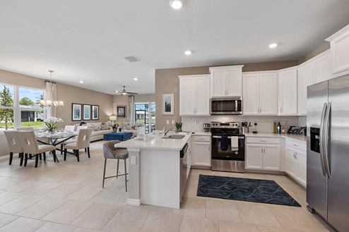 Inside of the Carrington model kitchen. Wood tile flooring and white walls. Kitchen has white countertops and cabinets with silver hardware, bar-style seating, and stainless-steel appliances. In the background the sliding glass door that leads to the back patio can be seen.