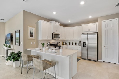 Inside of the Carrington model kitchen. Wood tile flooring and white walls. Kitchen has white countertops and cabinets with silver hardware, bar-style seating, and stainless-steel appliances.