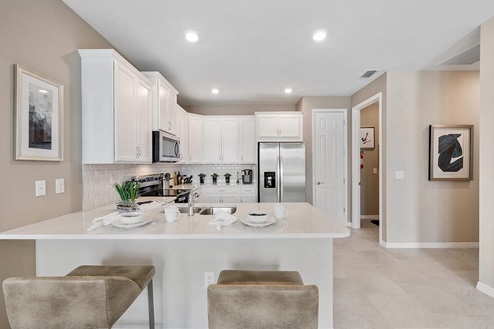 Inside of the Carrington model kitchen. Wood tile flooring and white walls. Kitchen has white countertops and cabinets with silver hardware, bar-style seating, and stainless-steel appliances.