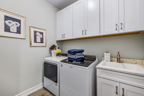 Inside of the Wheaton model laundry room. Room contains a white washer and dryer. Sink with cabinets and cabinets over the washer and dryer.