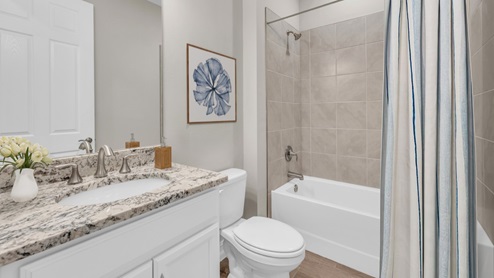 Inside of the Delray B model bathroom. This bathroom has a creme countertop with white cabinets and silver hardware. Shower-tub combo has grey tile and white tub. White toilet between vanity and shower-tub combo.