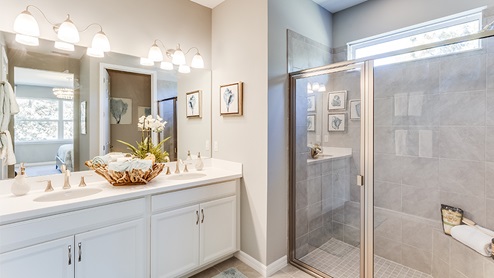 Inside of the Jaden model bathroom. Wood tile flooring, off-white walls, double vanity with white countertops and cabinets with silver hardware. Shower has grey tile and silver hardware.