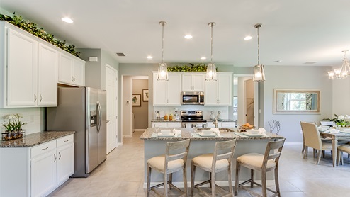 Inside of the Jaden model kitchen. Tile floor with teal and off-white walls. White cabinets with silver hardware. Kitchen and kitchen island uses grey countertops and bar style-seating.