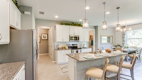Inside of the Jaden model kitchen. Tile floor with teal and off-white walls. White cabinets with silver hardware. Kitchen and kitchen island uses grey countertops and bar style-seating. In the background the dining room can be seen.