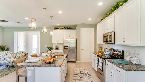 Inside of the Jaden model kitchen. Tile floor with teal and off-white walls. White cabinets with silver hardware. Kitchen and kitchen island uses grey countertops and bar style-seating. In the background the dining room can be seen.