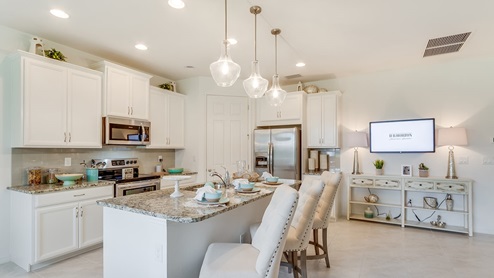 Inside of the Kellen model kitchen. Tile flooring and white walls. Kitchen has white cabinets and brown countertops with silver hardware. Kitchen island has bar-style seating.