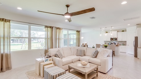 Inside of the Kellen model living room. Tile flooring and white walls. Living room has a couch, side table, two ottomen, and a coffee table. In the background you can see the kitchen area.