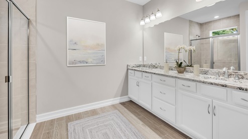 Inside of the Delray B model bathroom. Wood tile flooring with grey walls. Double vanity with grey countertops and white cabinets with silver hardware. Room features a shower with creme tile and glass shower door.