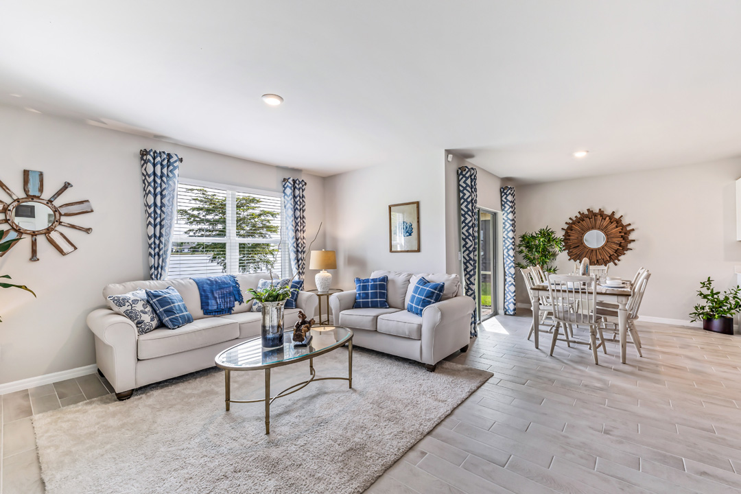 Inside of the Cali model living room. Grey wood tile floors and off-white walls. Room contains two small couches and a coffee table. The dining room can be seen in the background as well as the door to the outdoor patio. Room also contains a large window.