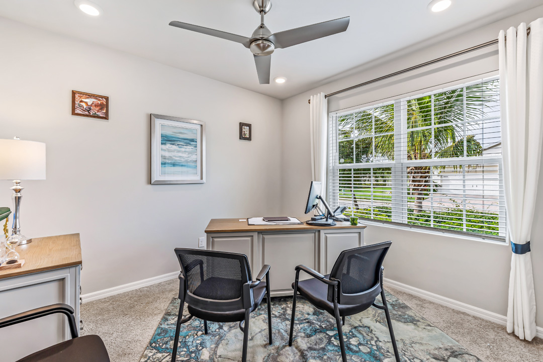 Inside of the Cali model den. Creme carpet with off-white walls. The room contains a desk with a laptop on it, two armchairs, and a small drawer. Room also has a large window.