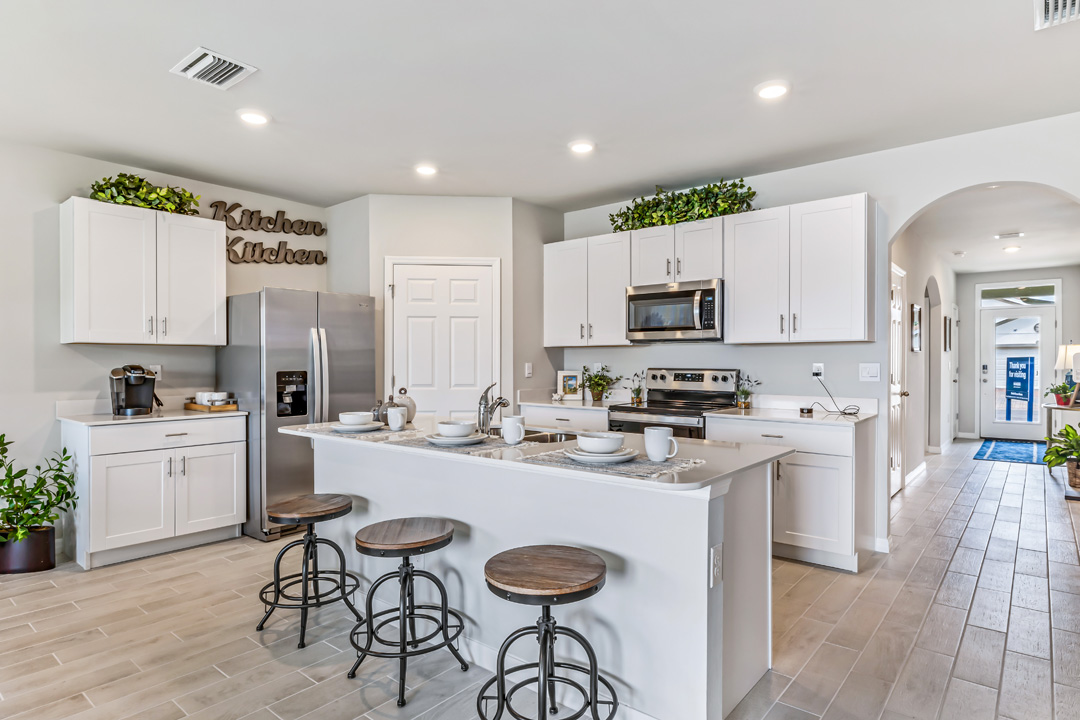Inside of the Cali model kitchen. Grey wood tile flooring and off-white walls. Kitchen has white cabinets with silver hardware, white countertops, and stainless-steel appliances. There is also a kitchen island with bar-style seating. In the background there is the hallway that connects to the front door and other rooms.