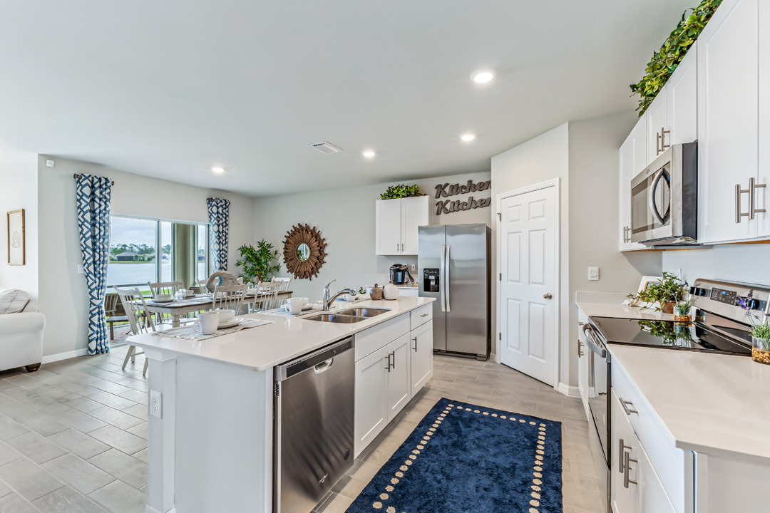 Inside of the Cali model kitchen. This angle shows the dishwasher on the inside of the kitchen island as well as the other stainless-steel appliances such as a refrigerator, stove, and microwave. In the background the dining room and back patio can be seen.