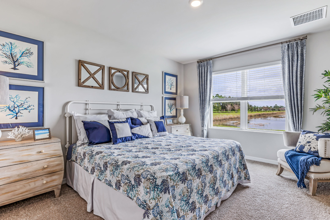 Inside of the Cali model bedroom one. Creme carpet with off-white walls. Room contains a large bed with dressers on either side. There is an armchair in the corner next to the room's window.